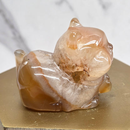 An Agate cat carving, with varying colors of orange, grey and white, on a wood stand against a white background