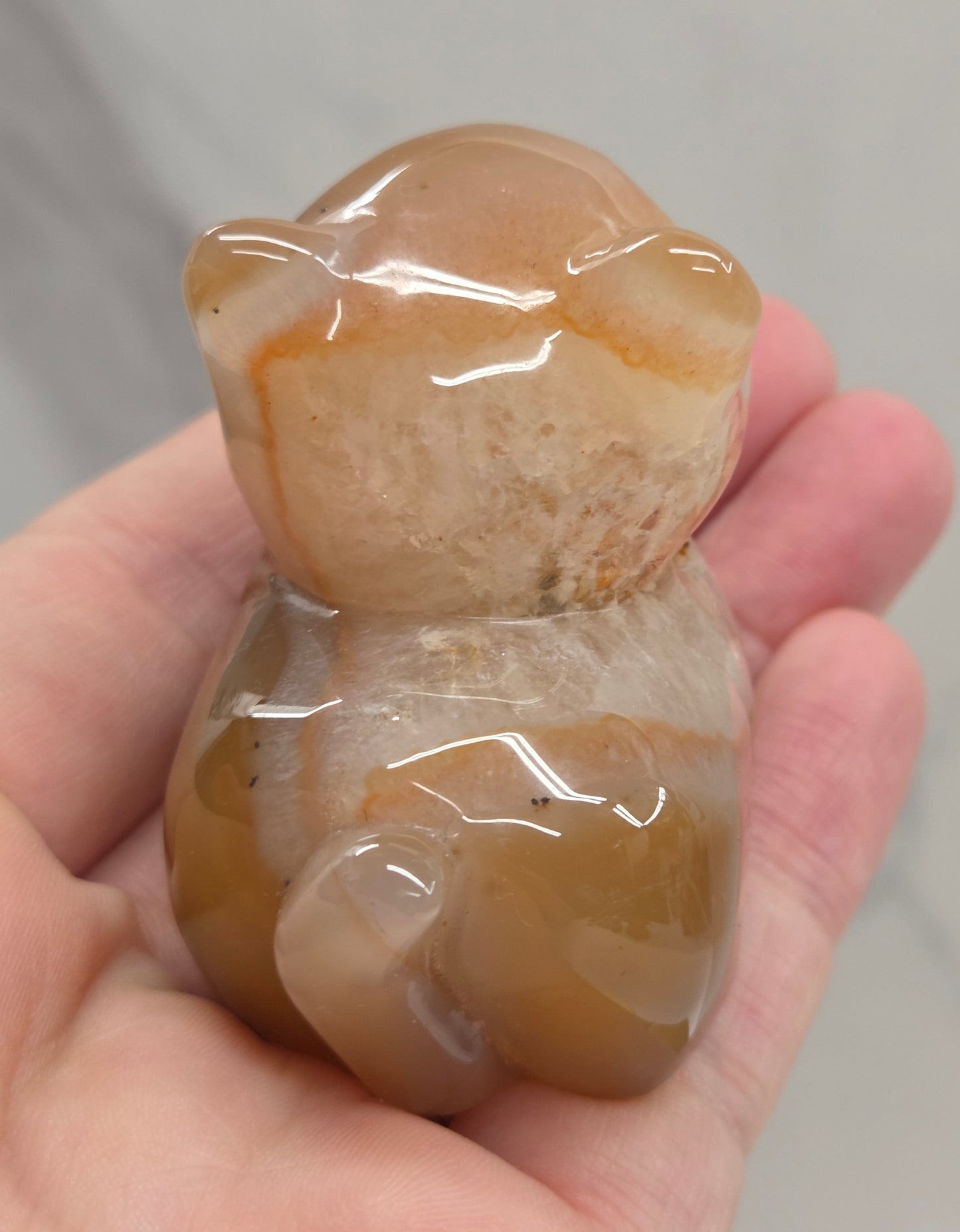 An Agate cat carving, with varying colors of orange, grey and white, being held by a hand against a white background