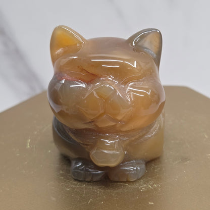 An Agate cat carving, with varying colors of orange, grey and white, on a wood stand against a white background