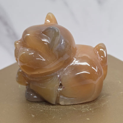 An Agate cat carving, with varying colors of orange, grey and white, on a wood stand against a white background