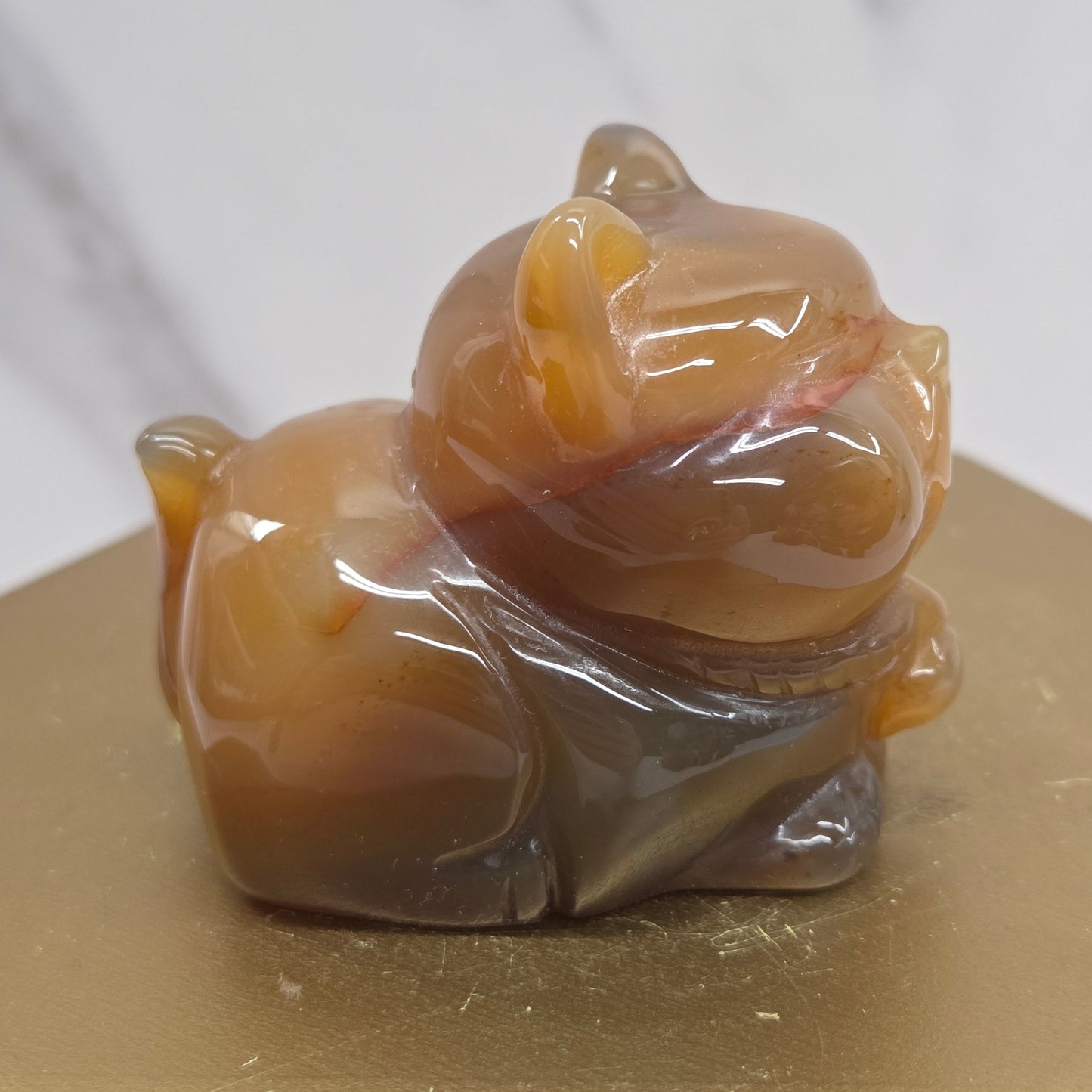 An Agate cat carving, with varying colors of orange, grey and white, on a wood stand against a white background