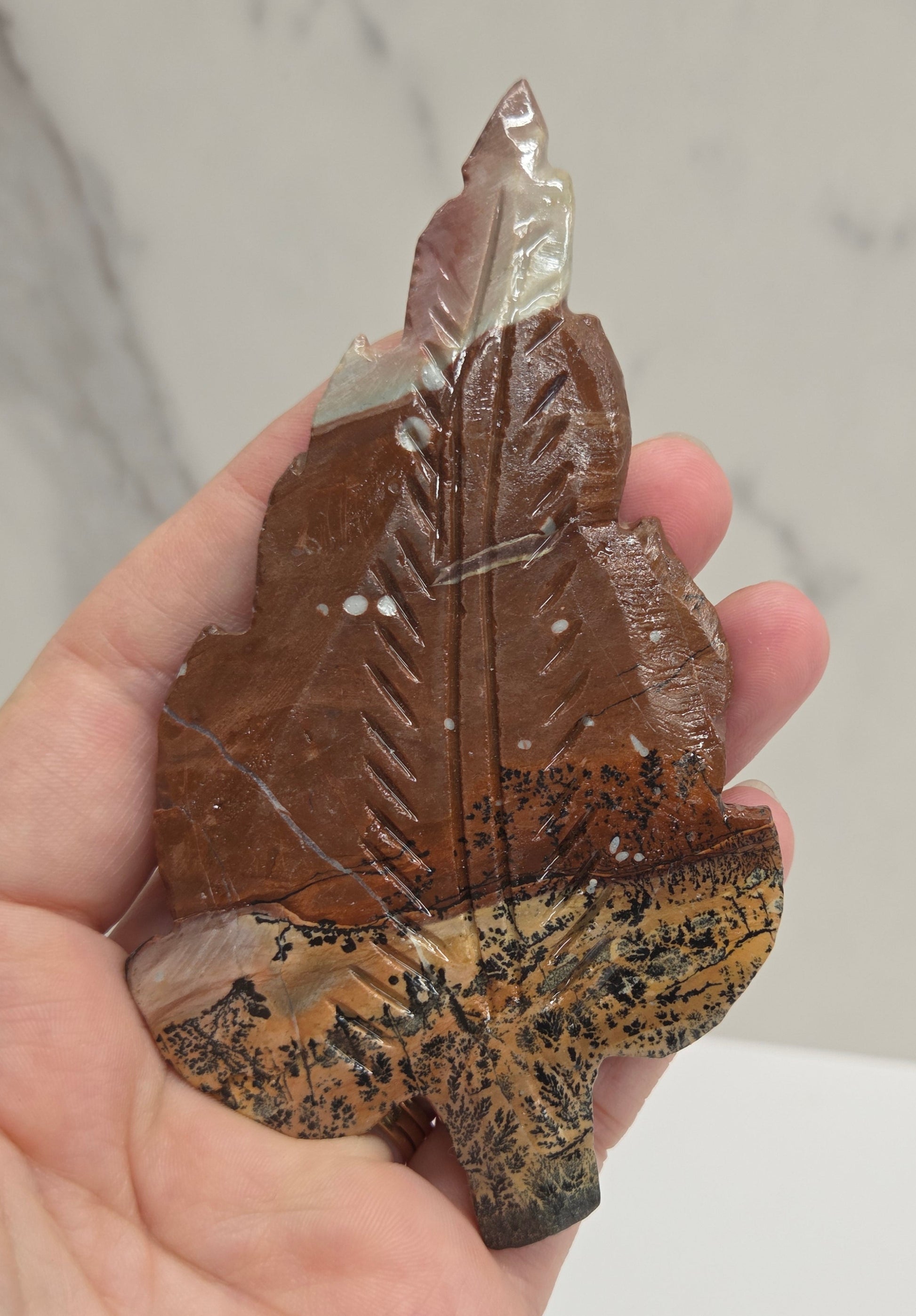 Hand holding a Picture Jasper leaf carving with black, gold, and white patterns against a neutral background