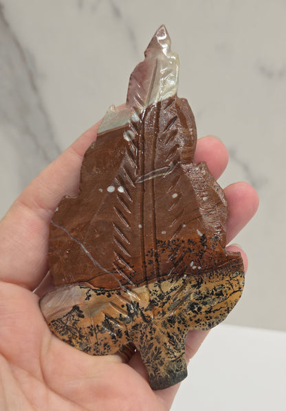Hand holding a Picture Jasper leaf carving with black, gold, and white patterns against a neutral background