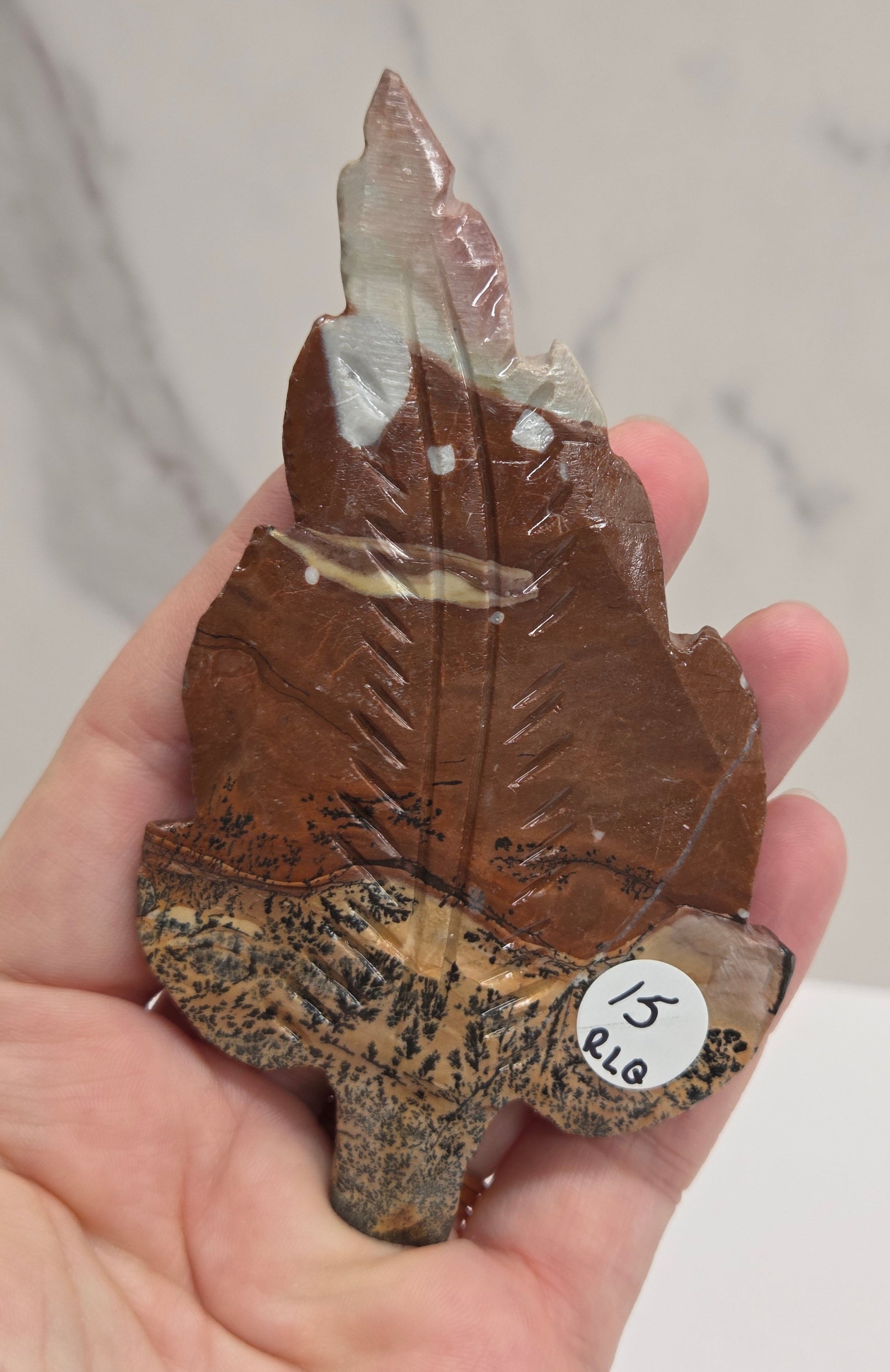 Hand holding a Picture Jasper leaf carving with black, gold, and white patterns against a neutral background