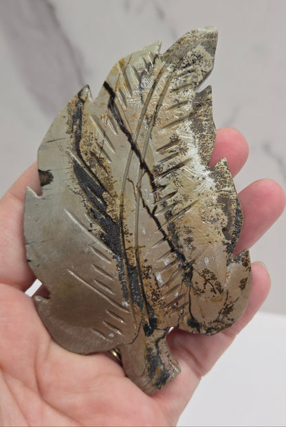 Hand holding a Picture Jasper leaf carving with black, gold, and white patterns against a neutral background
