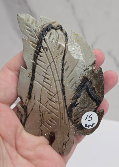 Hand holding a Picture Jasper leaf carving with black, gold, and white patterns against a neutral background