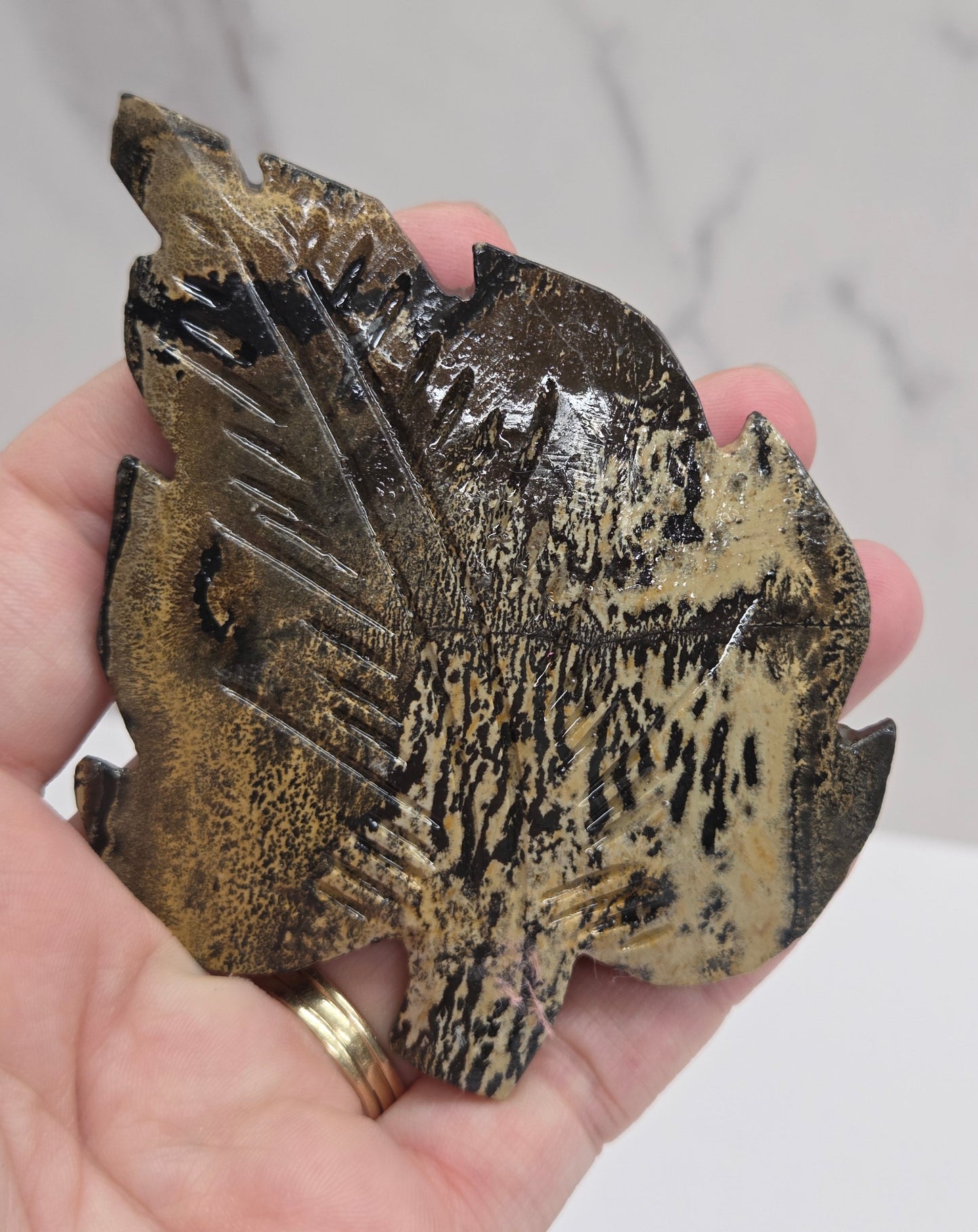 Hand holding a Picture Jasper leaf carving with black, gold, and white patterns against a neutral background