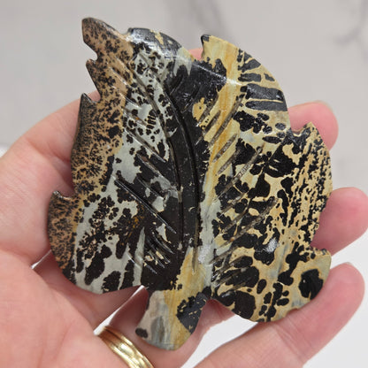 Hand holding a Picture Jasper leaf carving with black, gold, and white patterns against a neutral background