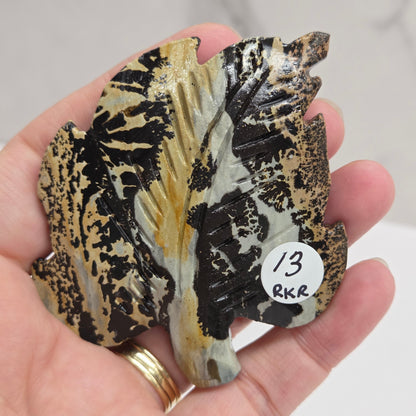 Hand holding a Picture Jasper leaf carving with black, gold, and white patterns against a neutral background