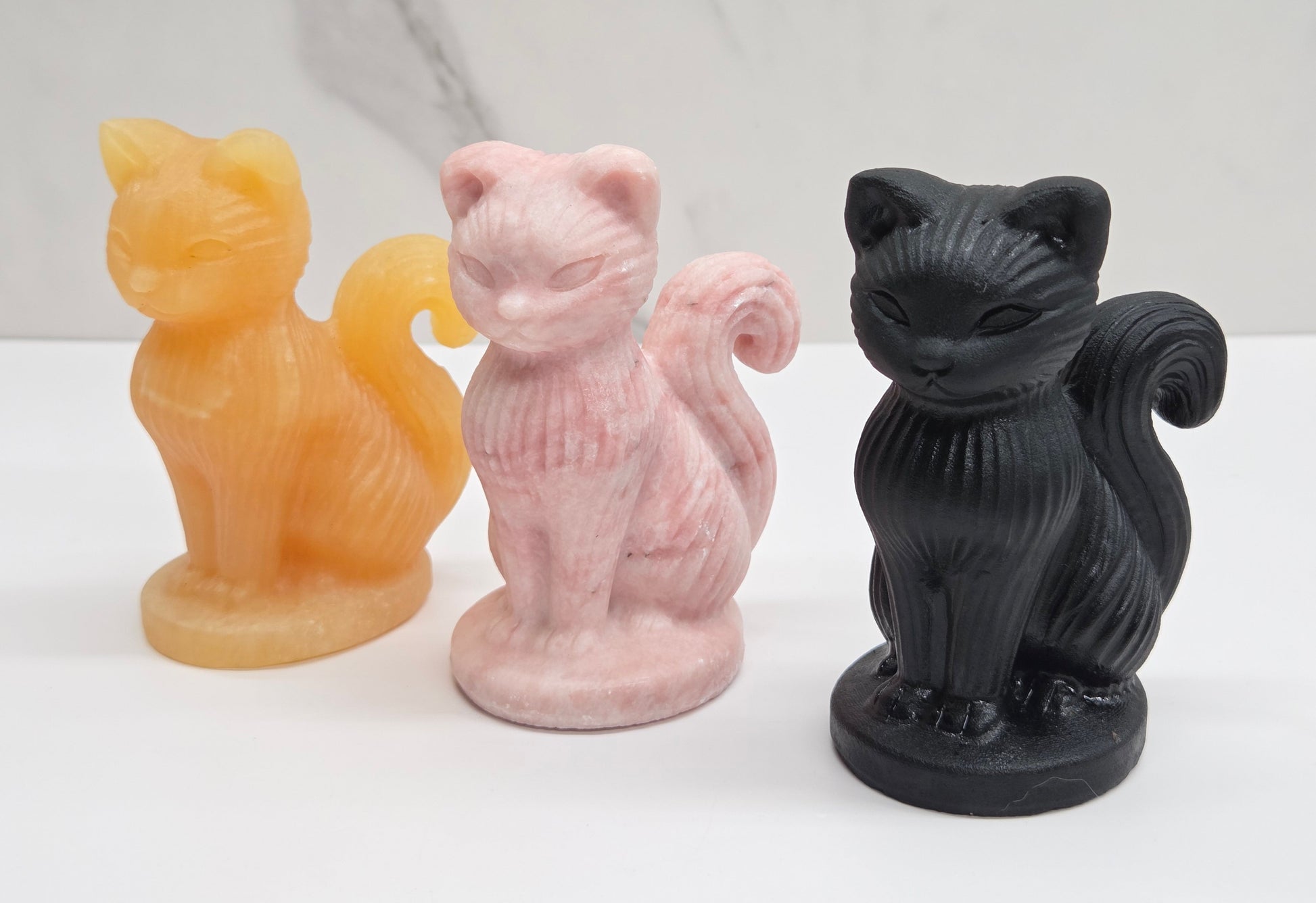 Three cat-shaped candles in orange, pink, and black on a white surface with a marble background.