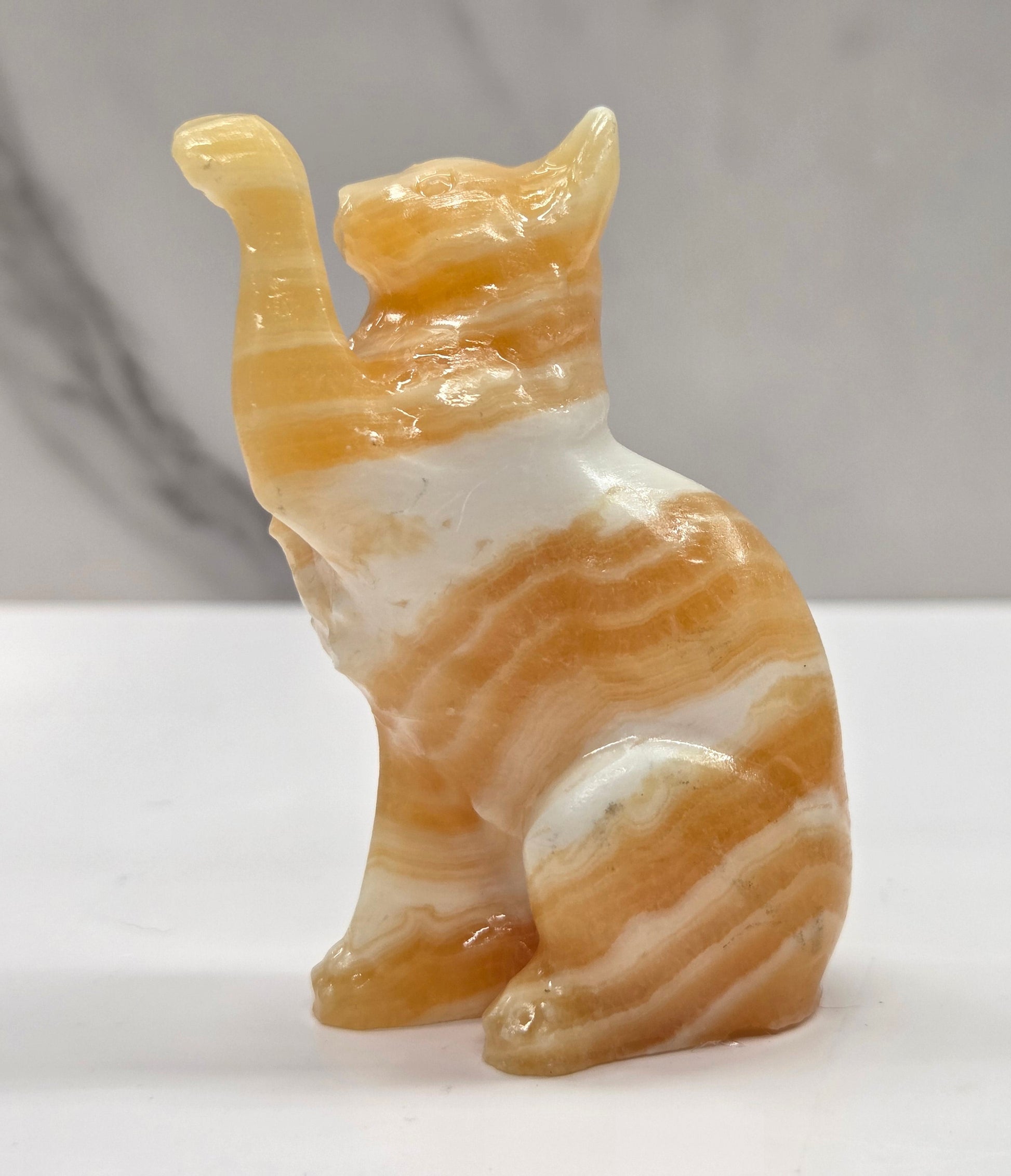 Cat crystal figurine made of  Orange Calcite, with banding of orange & white on a white surface with a light gray background