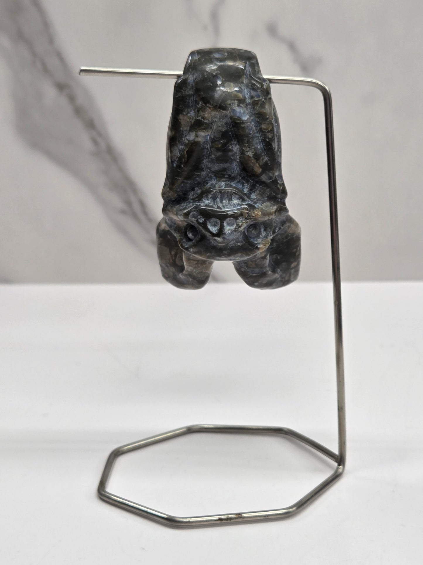 Que Sera bat carving with black and blue coloring, hanging from a metal stand, on a white background