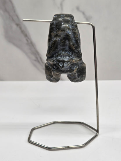 Que Sera bat carving with black and blue coloring, hanging from a metal stand, on a white background