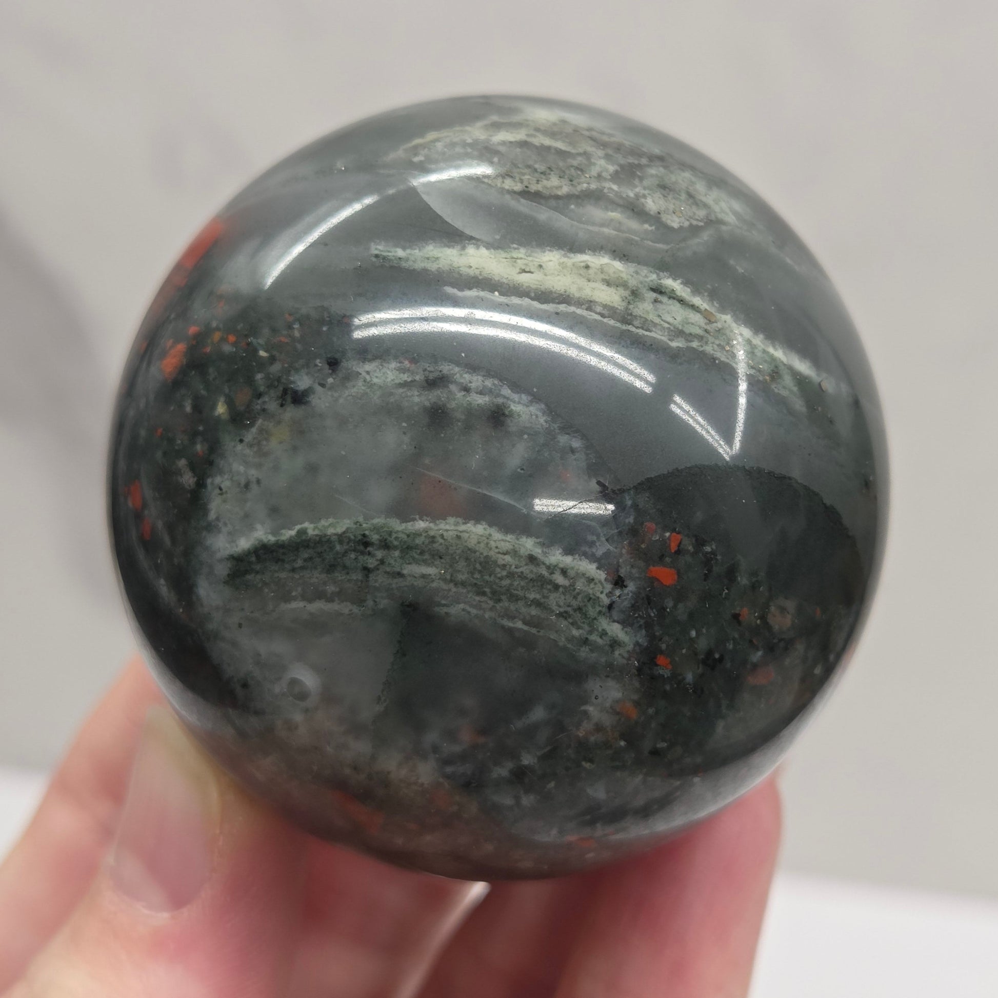 Hand holding an African Bloodstone sphere with black, grey, white and red coloring against a white background
