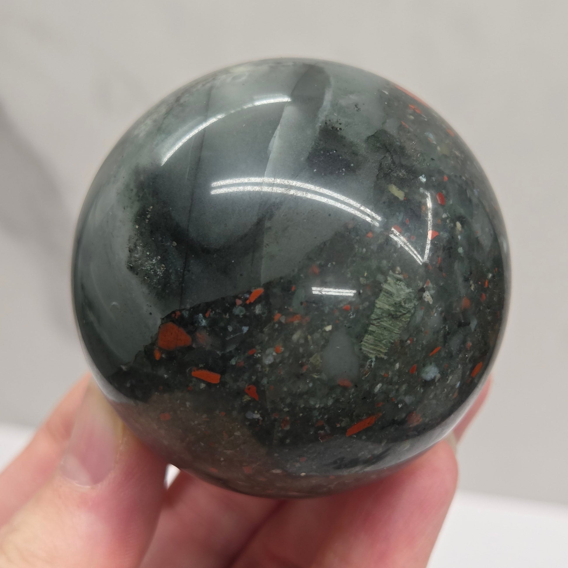 Hand holding an African Bloodstone sphere with black, grey, white and red coloring against a white background