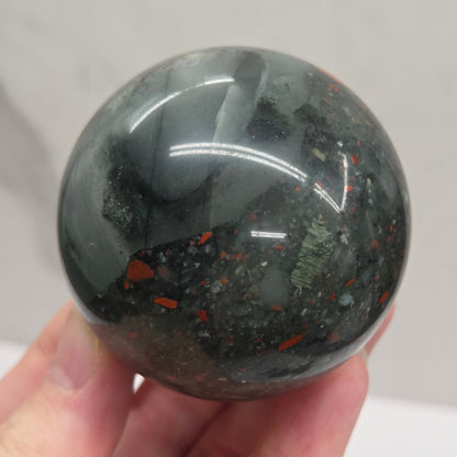 Hand holding an African Bloodstone sphere with black, grey, white and red coloring against a white background