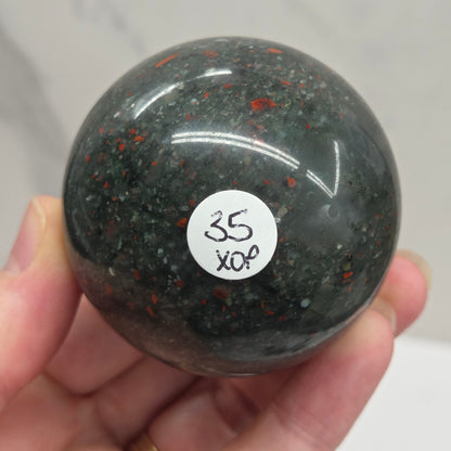 Hand holding an African Bloodstone sphere with black, grey, white and red coloring against a white background