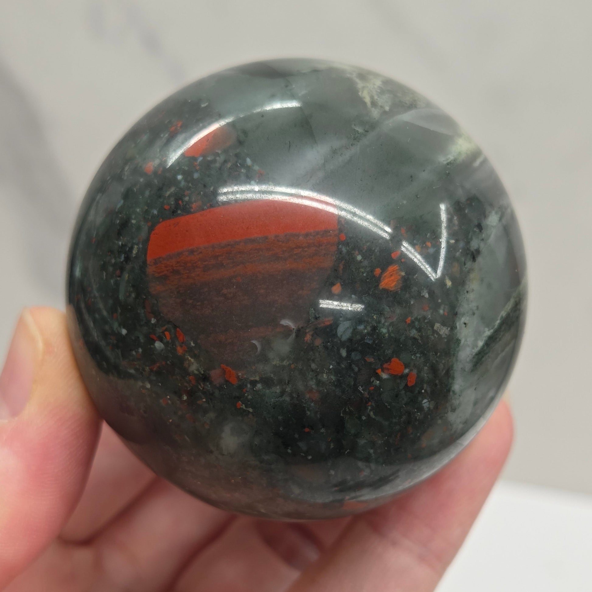 Hand holding an African Bloodstone sphere with black, grey, white and red coloring against a white background