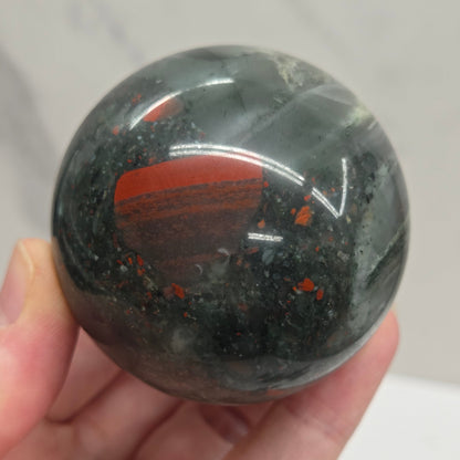 Hand holding an African Bloodstone sphere with black, grey, white and red coloring against a white background