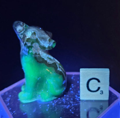 Volcano Agate wolf carving on a clear stand, next to a wooden block with 'C' under UV light