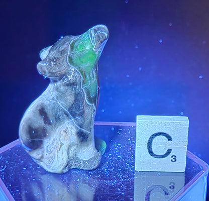 Volcano Agate wolf carving on a clear stand, next to a wooden block with 'C' under UV light