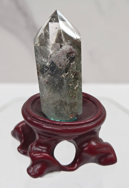 A clear Garden Quartz crystal with inclusions, mounted on a wooden stand with a circular cut-out at the base.