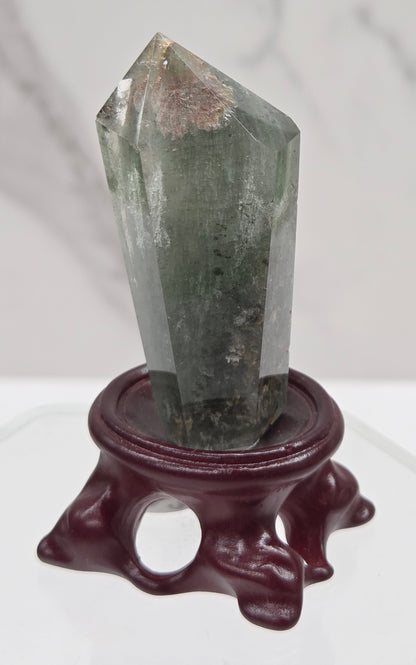 A clear Garden Quartz crystal with inclusions, mounted on a wooden stand with a circular cut-out at the base.