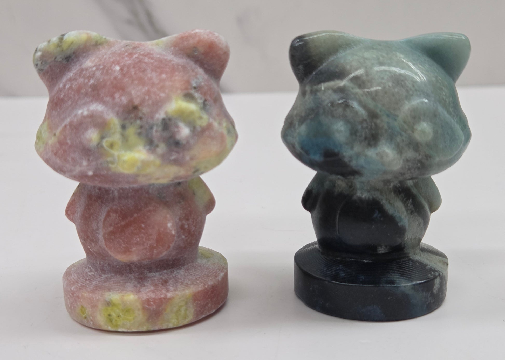 Two crystal raccoon carvings in a variety of colors and materials on a white surface with a neutral background