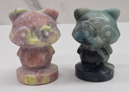 Two crystal raccoon carvings in a variety of colors and materials on a white surface with a neutral background