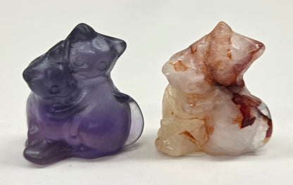 Two crystal cat figurines, one purple and one brown, on a white surface.