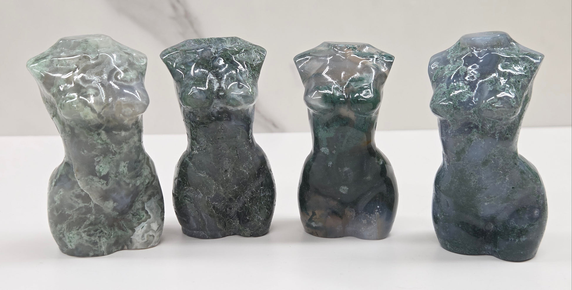 Four moss agate carvings of abstract human figures on a white surface with a marble-patterned wall in the background.
