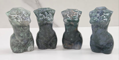 Four moss agate carvings of abstract human figures on a white surface with a marble-patterned wall in the background.