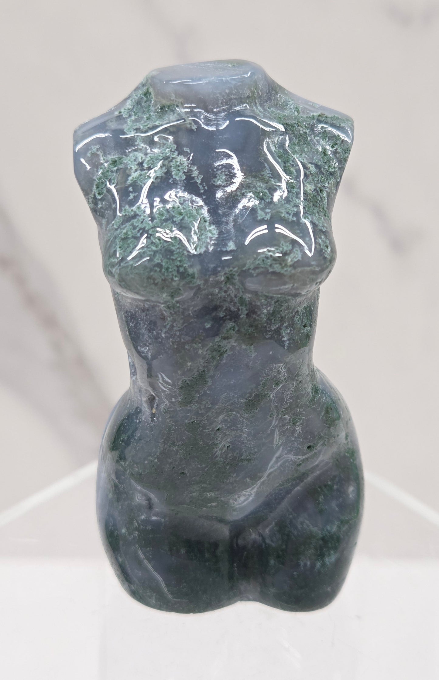 Moss agate carving of abstract human figures on a white surface with a marble-patterned wall in the background.