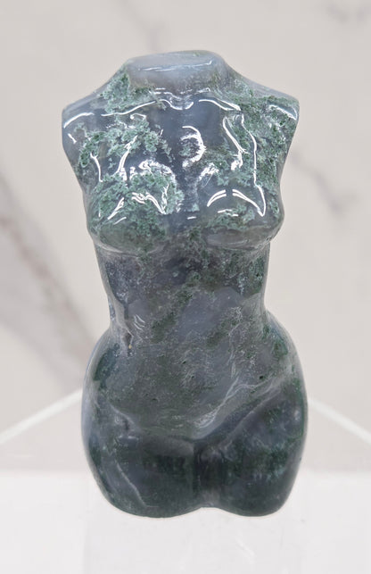 Moss agate carving of abstract human figures on a white surface with a marble-patterned wall in the background.