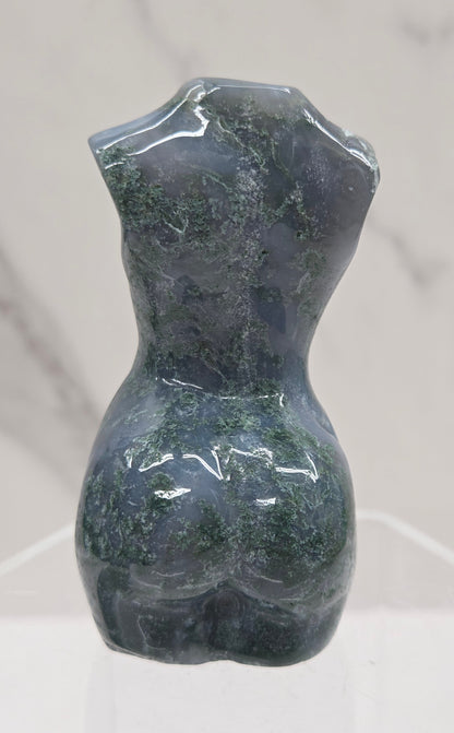 Moss agate carving of abstract human figures on a white surface with a marble-patterned wall in the background.