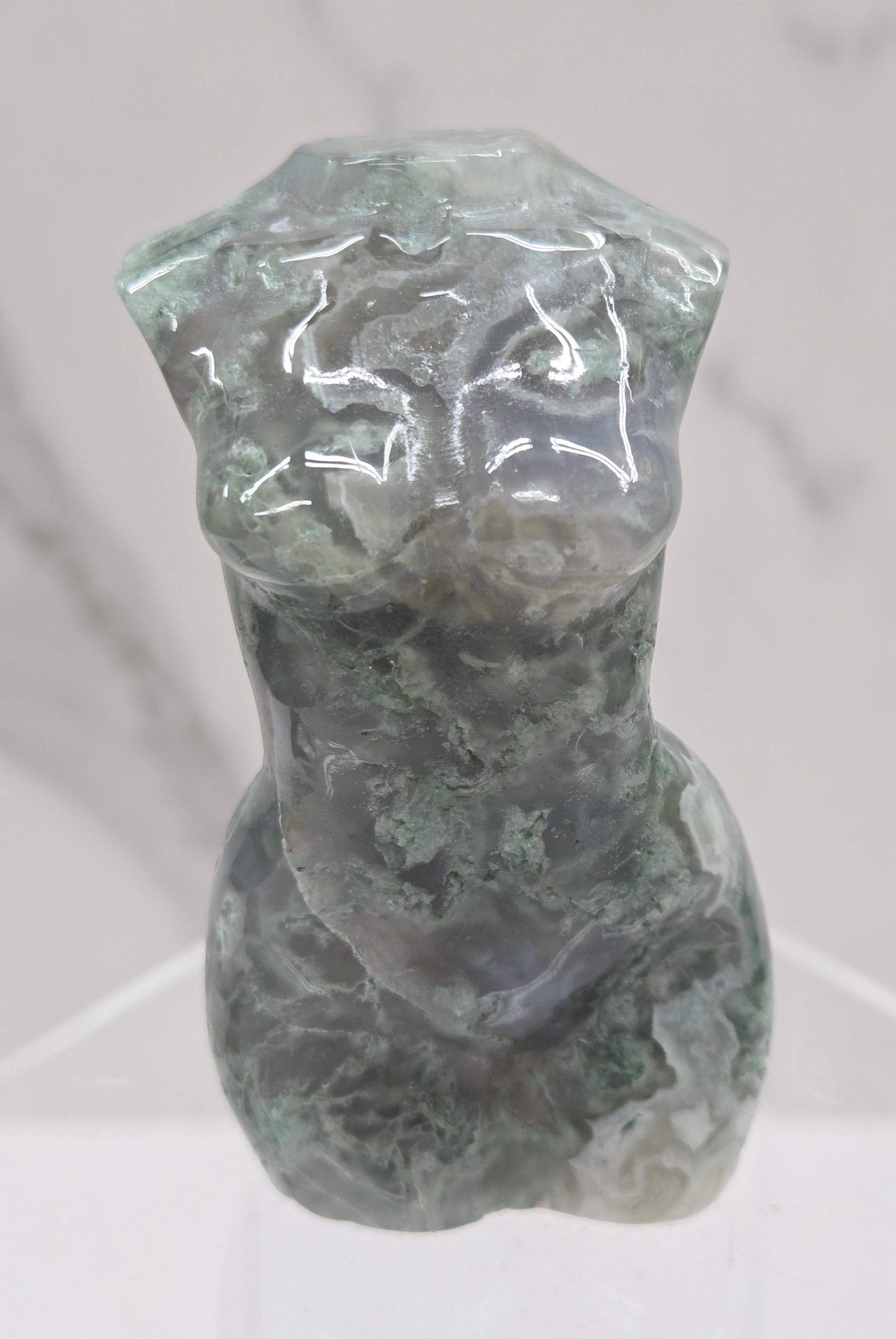 Moss agate carving of abstract human figures on a white surface with a marble-patterned wall in the background.