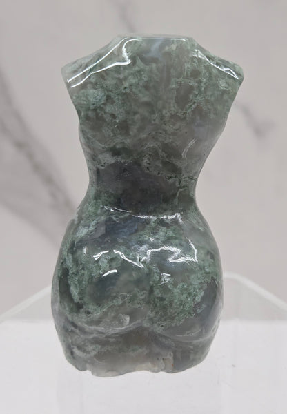 Moss agate carving of abstract human figures on a white surface with a marble-patterned wall in the background.
