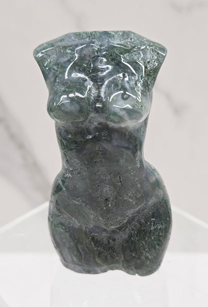 Moss agate carving of abstract human figures on a white surface with a marble-patterned wall in the background.