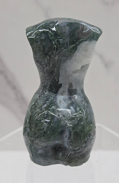 Moss agate carving of abstract human figures on a white surface with a marble-patterned wall in the background.