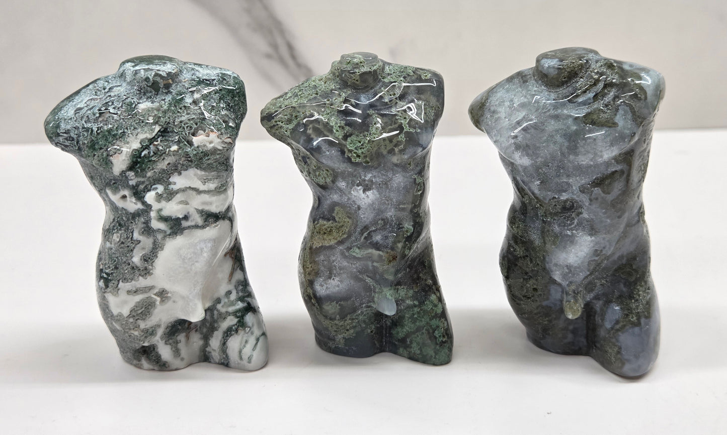 Three moss agate sculptures of human torsos on a white surface with a neutral background