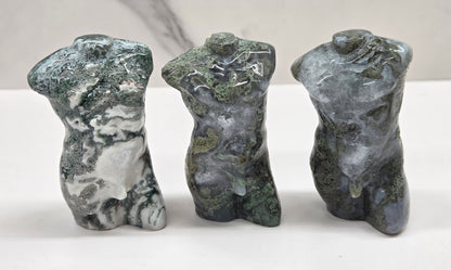 Three moss agate sculptures of human torsos on a white surface with a neutral background