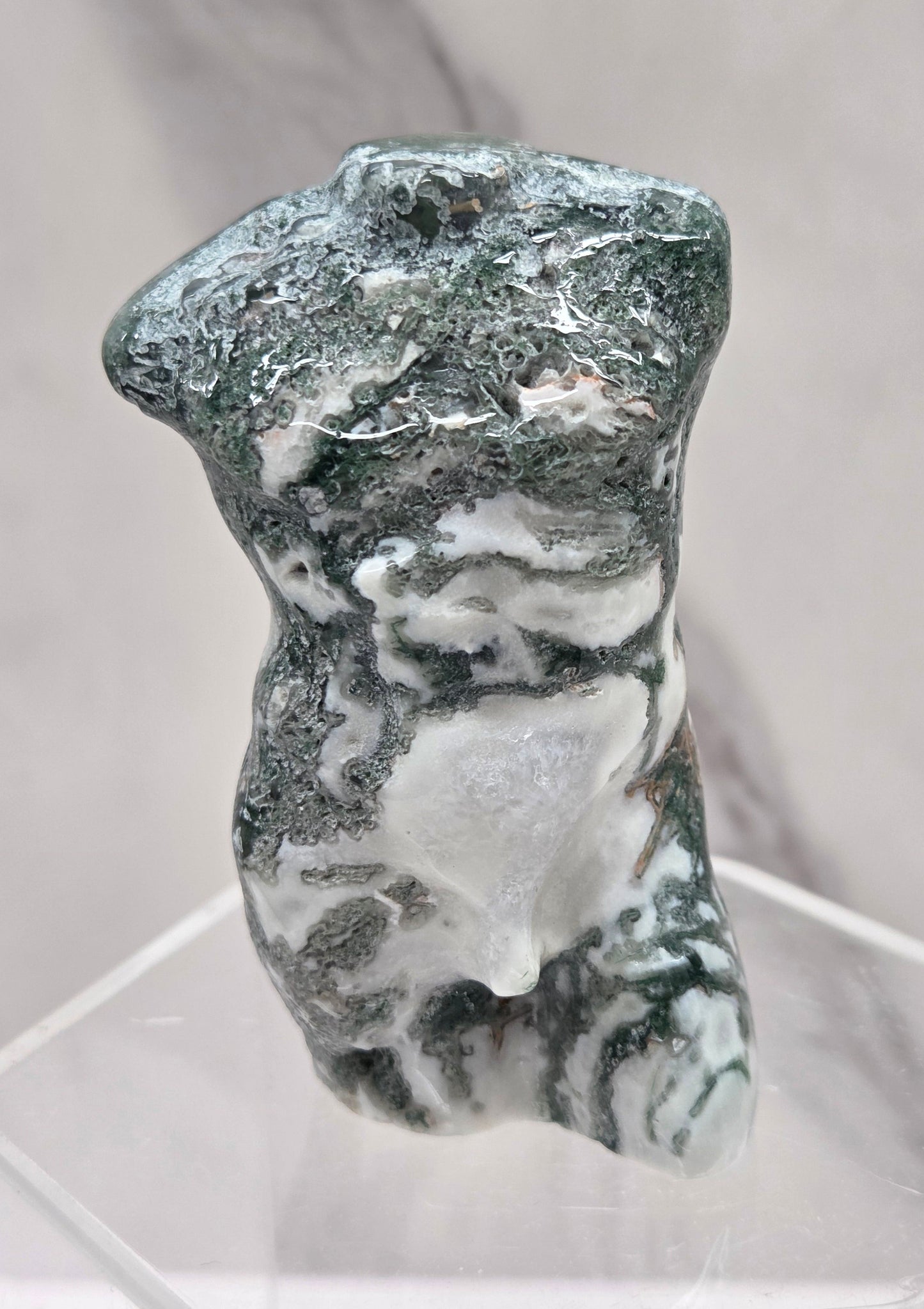 Moss agate carving of abstract human figures on a white surface with a marble-patterned wall in the background.