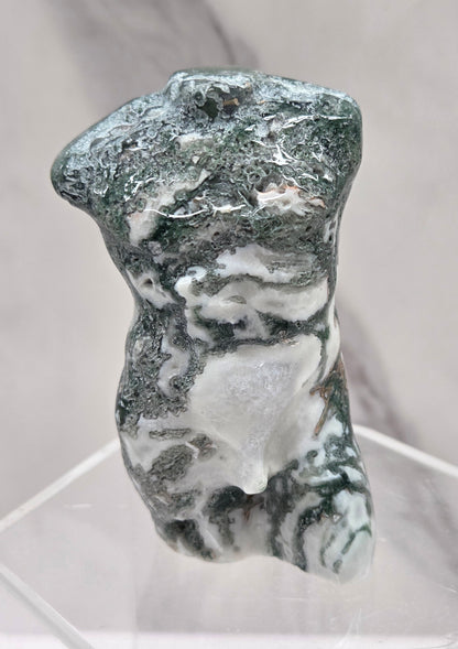 Moss agate carving of abstract human figures on a white surface with a marble-patterned wall in the background.