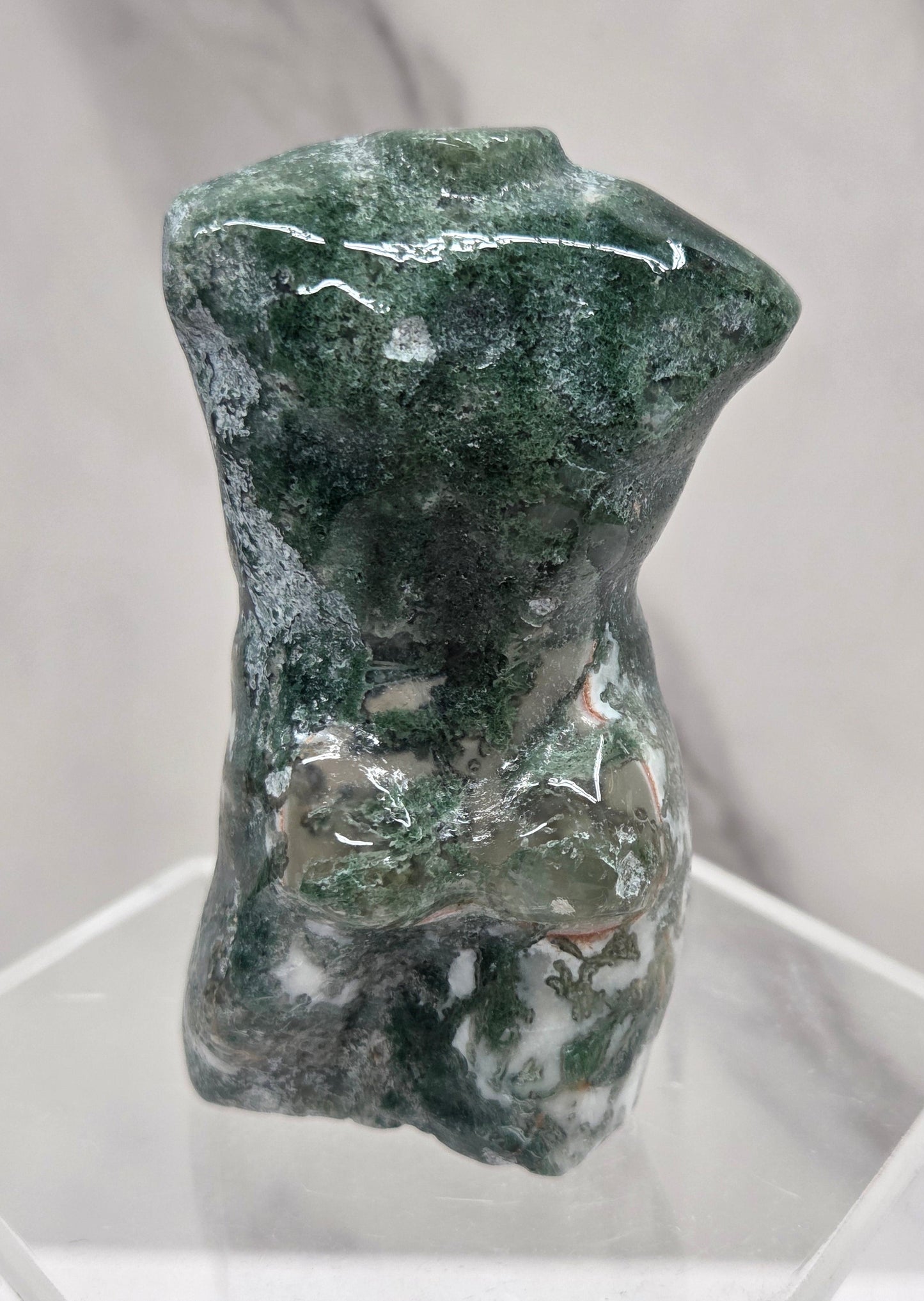 Moss agate carving of abstract human figures on a white surface with a marble-patterned wall in the background.