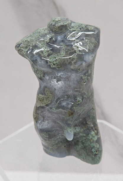 Moss agate carving of abstract human figures on a white surface with a marble-patterned wall in the background.