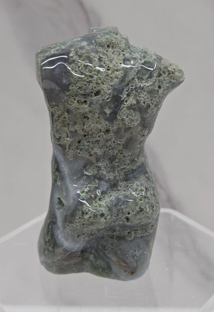 Moss agate carving of abstract human figures on a white surface with a marble-patterned wall in the background.
