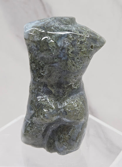 Moss agate carving of abstract human figures on a white surface with a marble-patterned wall in the background.