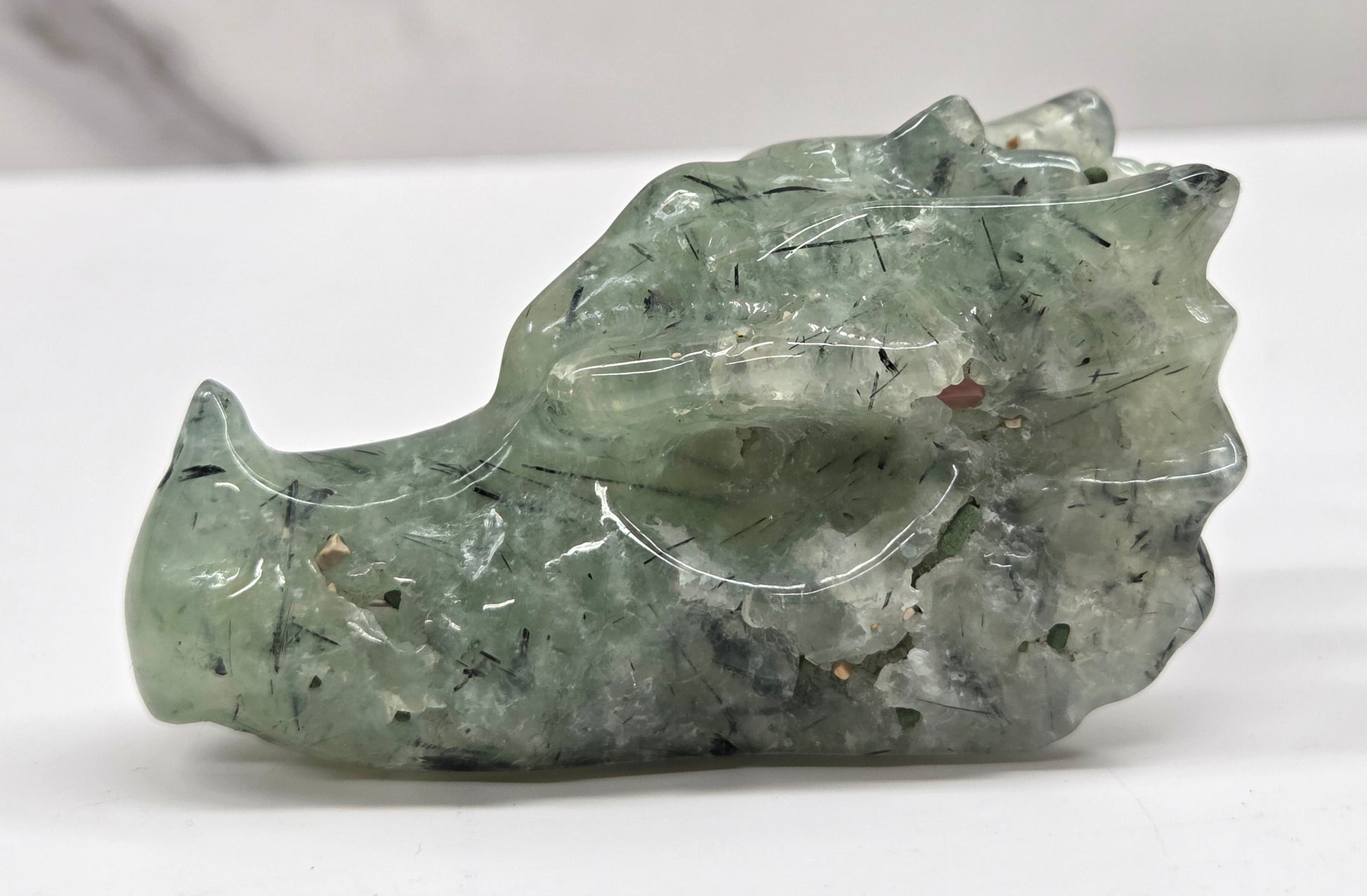 Green Prehnite dragon head carving with black inclusions on a white background