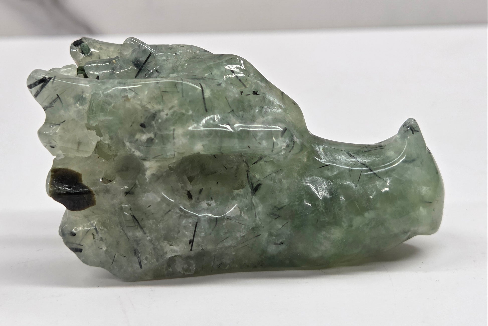 Green Prehnite dragon head carving with black inclusions on a white background
