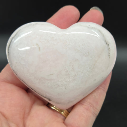 Heart-shaped pale pink calcite carving held in a hand against a dark background